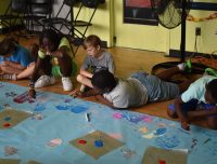 Children working on an art project at summer camp, adding colorful creatures to a long blue backdrop with paints and markers