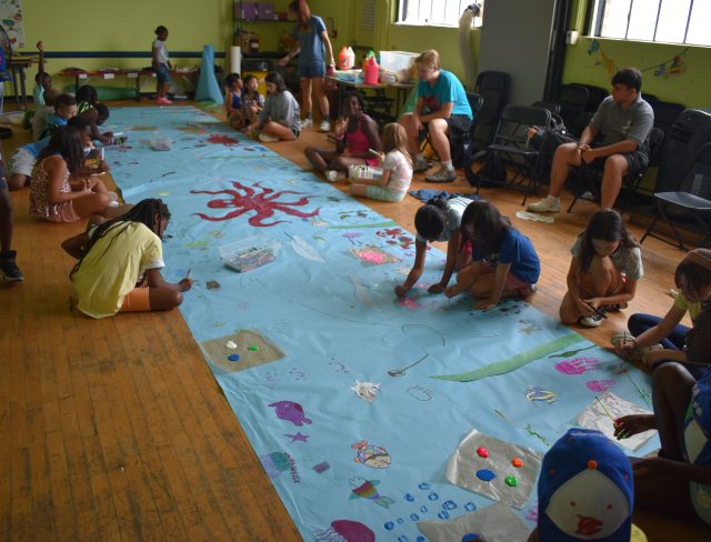 A group of children working on background for an art project, the long blue paper is being decorated with painted and stamped undersea creatures