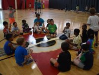 youn kids playing Duck Duck Goose at Summer Camp with an adult smiling at them.
