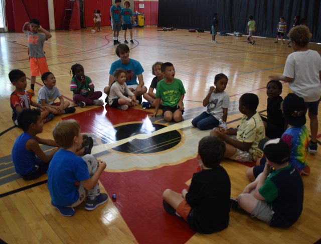 youn kids playing Duck Duck Goose at Summer Camp with an adult smiling at them.