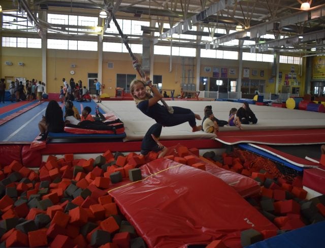 a girl with blonde hair swings on a rope over a foam pit, smiling while enjoying the aviator camp facilities