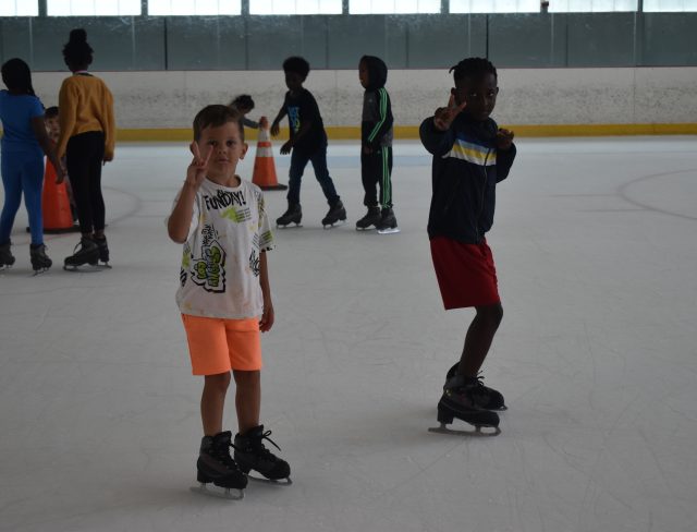Young boys wearing ice skates, shorts, and sweatshirts glide by while throwing "peace signs" to the camera