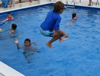A camper jumping into one of the pools at summer camp while another waves at the camera from the water