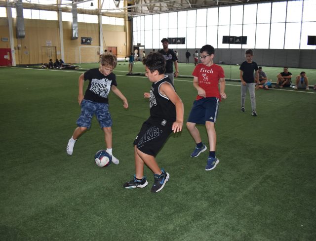 Three young campers playing soccer on the turf fields, wearing t-shirts and shorts while dribbling a ball together