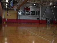 An empty indoor basketball gymnasium with a polished wooden floor, red and black padded walls, and a basketball hoop. The gym is well-lit with overhead lights and features a scoreboard on the far wall above viewing windows. Benches are aligned along the side.