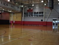 A spacious indoor basketball court with polished wooden floors, red and beige walls, and a view of the audience stands on the upper level. There's a backboard and hoop on the right side of the image.