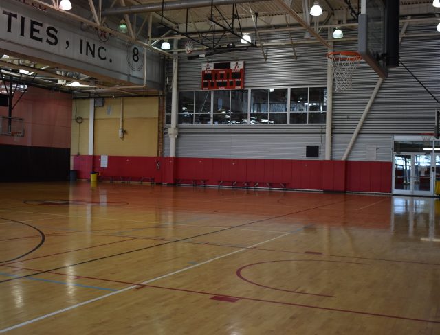 A spacious indoor basketball court with polished wooden floors, red and beige walls, and a view of the audience stands on the upper level. There's a backboard and hoop on the right side of the image.