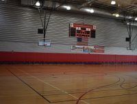 An empty gymnasium with wooden flooring, two basketball hoops, and a large scoreboard displaying a score of 0-0.