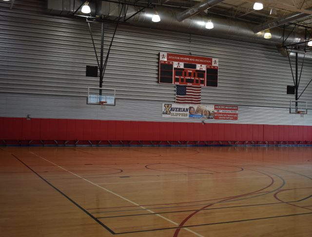 An empty gymnasium with wooden flooring, two basketball hoops, and a large scoreboard displaying a score of 0-0.
