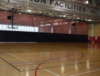 A spacious indoor sports facility with a polished wooden basketball court, multiple basketball hoops, and a red divider curtain. Overhead, a sign reads "ON FACILITIES, INC." Windows at the back provide natural light, and the ceiling is equipped with lights and ductwork.