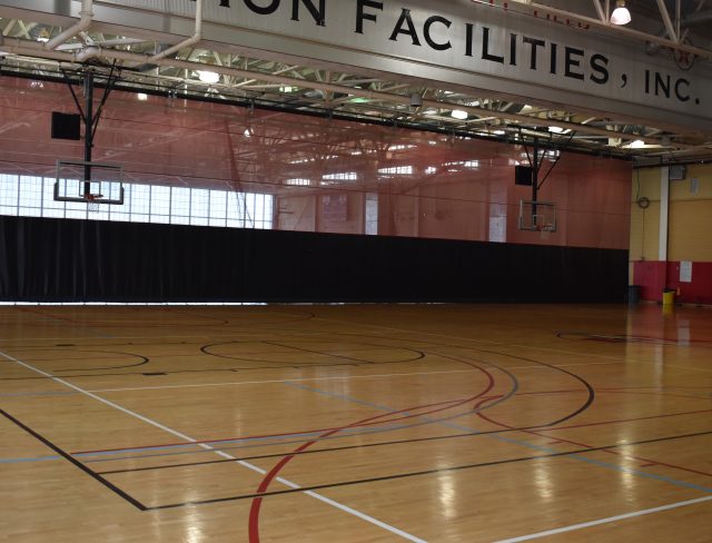 A spacious indoor sports facility with a polished wooden basketball court, multiple basketball hoops, and a red divider curtain. Overhead, a sign reads "ON FACILITIES, INC." Windows at the back provide natural light, and the ceiling is equipped with lights and ductwork.