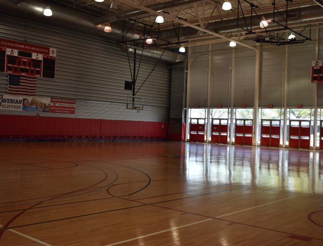 A well-lit indoor basketball court with polished wooden floors, red padding along the walls, and an electronic scoreboard displaying "00." There are multiple basketball hoops and large windows with red frames lining the side of the court.