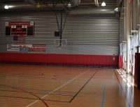 An empty indoor basketball court with wooden flooring and red wall padding. A large electronic scoreboard and American flag are mounted on the wall. Sunlight streams in through the windows on the right side. Banners are displayed on the wall.