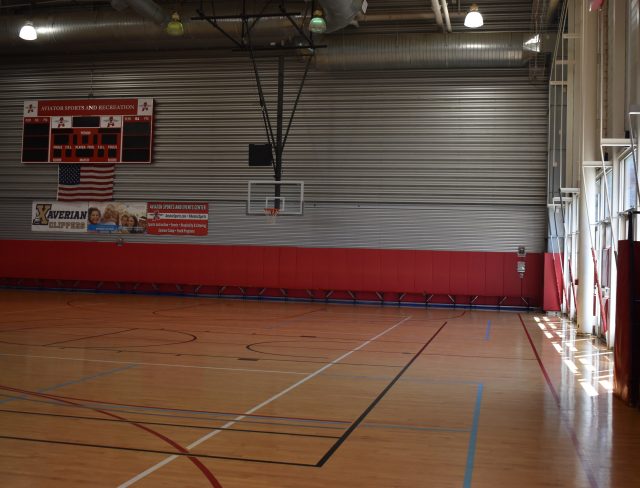An empty indoor basketball court with wooden flooring and red wall padding. A large electronic scoreboard and American flag are mounted on the wall. Sunlight streams in through the windows on the right side. Banners are displayed on the wall.