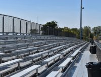 A row of empty metal bleachers under a clear blue sky, with trash cans placed along a chain-link fence to the right. Trees and a sports field are visible in the background.