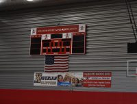 An indoor sports facility features a scoreboard with the label "Aviator Sports and Recreation." Below the scoreboard is an American flag and advertisements, including one for "Xaverian Clippers." A basketball hoop is mounted on the right side.