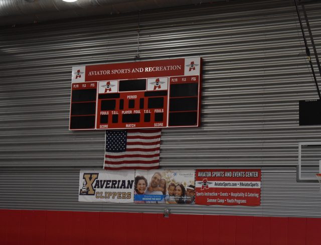 An indoor sports facility features a scoreboard with the label "Aviator Sports and Recreation." Below the scoreboard is an American flag and advertisements, including one for "Xaverian Clippers." A basketball hoop is mounted on the right side.
