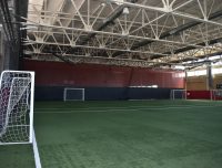 The image shows an indoor soccer field with artificial turf. There are three small goals visible, each with white netting, placed evenly along the back wall. The ceiling features metal trusses and large lights. The space is well-lit and spacious.