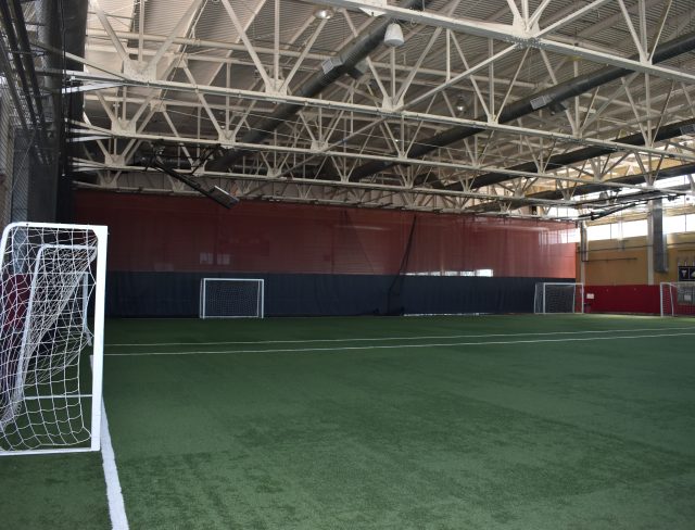 The image shows an indoor soccer field with artificial turf. There are three small goals visible, each with white netting, placed evenly along the back wall. The ceiling features metal trusses and large lights. The space is well-lit and spacious.