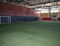Indoor soccer field with artificial grass and three soccer goals placed against red and yellow walls. The space has a high ceiling with visible beams and lighting fixtures.