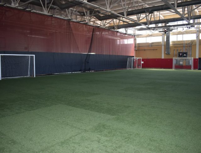 Indoor soccer field with artificial grass and three soccer goals placed against red and yellow walls. The space has a high ceiling with visible beams and lighting fixtures.