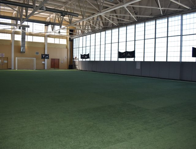 Indoor sports arena with artificial turf and high ceilings. A soccer goal is visible against a wall in the far corner. Large windows along one side let in natural light. Metal beams support the ceiling, and several flags are hung on the wall.