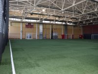 Show thumbnail preview An indoor sports facility with artificial turf covers the floor. Industrial-style ceiling with exposed beams. A scoreboard, utility doors, and a red railing run along the far wall. Small soccer goals are positioned on the right side of the space.