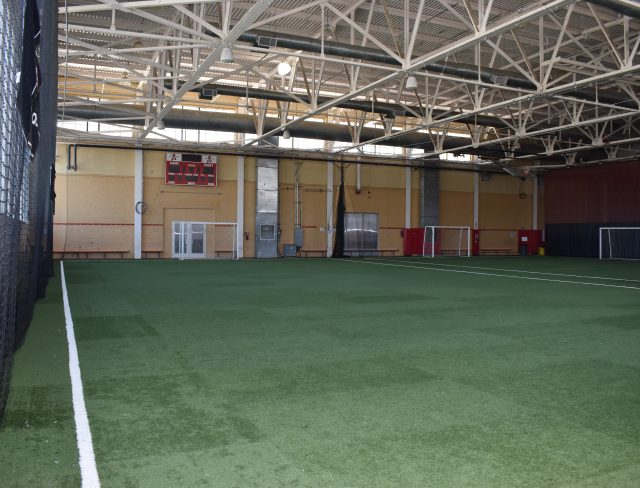 Show thumbnail preview An indoor sports facility with artificial turf covers the floor. Industrial-style ceiling with exposed beams. A scoreboard, utility doors, and a red railing run along the far wall. Small soccer goals are positioned on the right side of the space.