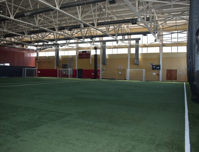 An indoor soccer field with artificial turf, surrounded by walls and a high ceiling with metal beams. Multiple goals are visible, along with windows letting in natural light. The area appears spacious and well-lit.