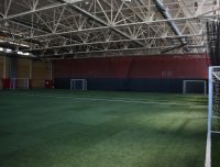 An indoor soccer field with artificial turf, white markings, and multiple goals. The ceiling features exposed beams and large ducts, and the walls are painted red and yellow. Nets hang along one side of the field, partially covering the area.