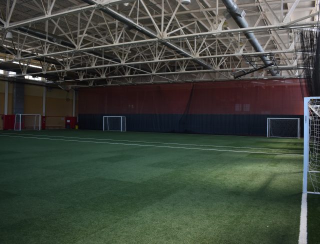 An indoor soccer field with artificial turf, white markings, and multiple goals. The ceiling features exposed beams and large ducts, and the walls are painted red and yellow. Nets hang along one side of the field, partially covering the area.