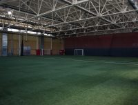 An indoor sports facility with a large artificial turf field. The area is spacious and well-lit with a metal truss ceiling. Several soccer goals and nets are positioned around the perimeter of the field. The walls are painted in neutral colors.