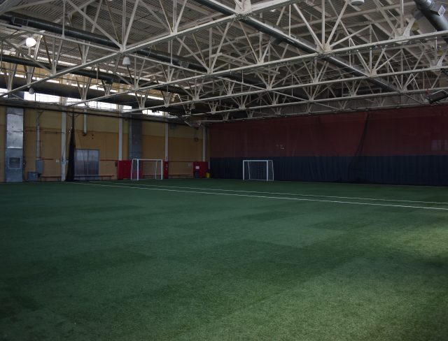 An indoor sports facility with a large artificial turf field. The area is spacious and well-lit with a metal truss ceiling. Several soccer goals and nets are positioned around the perimeter of the field. The walls are painted in neutral colors.