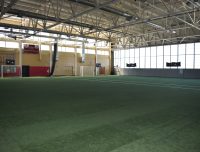 An indoor soccer field with artificial grass. The field has two goals at opposite ends and is enclosed by walls with large windows letting in natural light. The facility includes high ceilings, ventilation ducts, and a scoreboard mounted on one wall.