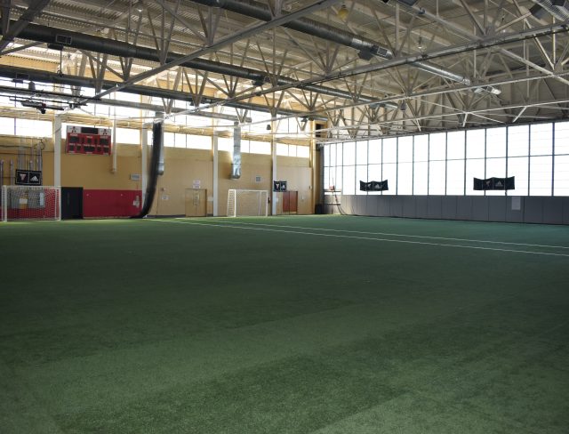 An indoor soccer field with artificial grass. The field has two goals at opposite ends and is enclosed by walls with large windows letting in natural light. The facility includes high ceilings, ventilation ducts, and a scoreboard mounted on one wall.