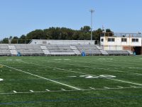 Show thumbnail preview A football field with visible yard lines marked in white. The field has artificial green turf and is surrounded by a metal fence. In the background, there are bleachers and a small building with an American flag on a pole, both under a clear blue sky.