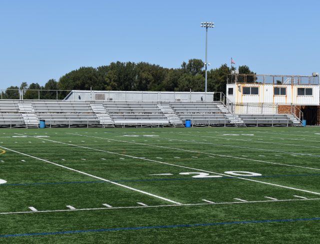 Show thumbnail preview A football field with visible yard lines marked in white. The field has artificial green turf and is surrounded by a metal fence. In the background, there are bleachers and a small building with an American flag on a pole, both under a clear blue sky.