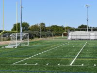 An empty outdoor sports field with goalposts, a soccer goal, and metal bleachers. The field has marked lines for various sports, and several light poles are visible in the background. Trees and a clear blue sky can be seen beyond the field.