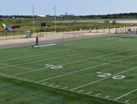 Show thumbnail preview A green artificial turf sports field with goalposts and white line markings. The field includes numbers for yard lines such as 10, 20. Beyond the field, there is a fence, some construction equipment, and a grassy area with structures in the distance.