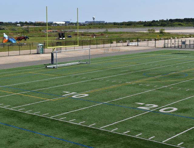 Show thumbnail preview A green artificial turf sports field with goalposts and white line markings. The field includes numbers for yard lines such as 10, 20. Beyond the field, there is a fence, some construction equipment, and a grassy area with structures in the distance.