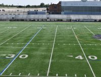 A wide view of an empty football field marked with yard lines, including the 30 and 40-yard lines in the foreground. Bleachers and a building with the name "Floyd Bennett Field" are visible in the background, situated under a clear sky.