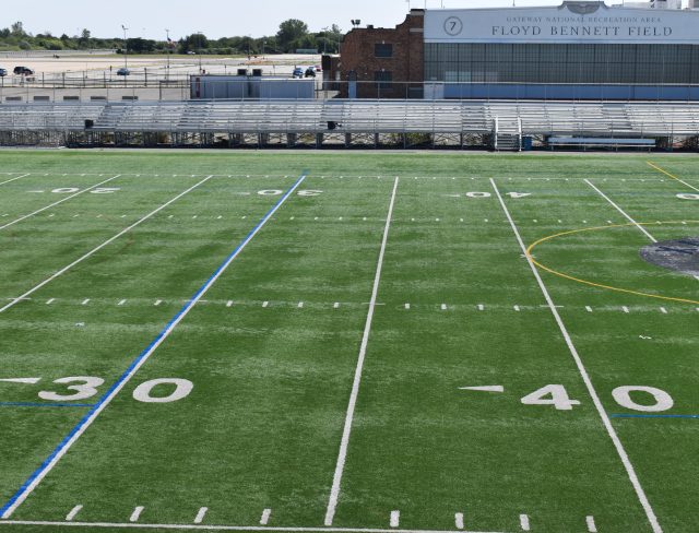A wide view of an empty football field marked with yard lines, including the 30 and 40-yard lines in the foreground. Bleachers and a building with the name "Floyd Bennett Field" are visible in the background, situated under a clear sky.