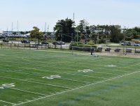 An empty sports field with green turf and white yard markings, including numbers 20, 25, 30, and 40. A single soccer goal is seen in the background. Beyond the field, there is a road with some parked cars, trees, and a view of boats docked at a marina.