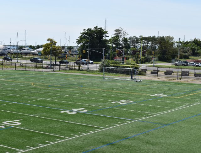 An empty sports field with green turf and white yard markings, including numbers 20, 25, 30, and 40. A single soccer goal is seen in the background. Beyond the field, there is a road with some parked cars, trees, and a view of boats docked at a marina.