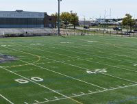 Show thumbnail preview A well-maintained football field with yard lines marked from 50 to 30. Behind the field are bleachers and a building labeled "Floyd Bennett Field." In the background, there are trees, a flagpole, and several boats docked near a water body.