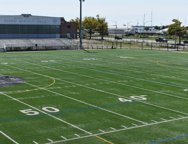 Show thumbnail preview A well-maintained football field with yard lines marked from 50 to 30. Behind the field are bleachers and a building labeled "Floyd Bennett Field." In the background, there are trees, a flagpole, and several boats docked near a water body.
