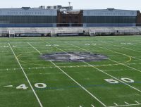 A wide view of the Floyd Bennett Field football stadium, featuring a large, well-maintained green turf field marked with yard lines and numbers. The stadium benches and the main building, labeled "Floyd Bennett Field," are visible in the background.