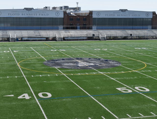 A wide view of the Floyd Bennett Field football stadium, featuring a large, well-maintained green turf field marked with yard lines and numbers. The stadium benches and the main building, labeled "Floyd Bennett Field," are visible in the background.