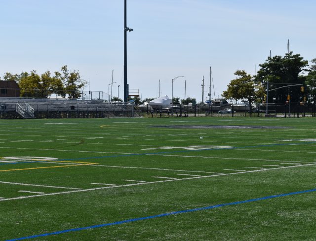 A green, grass-covered football field with white and yellow yard lines, and blue soccer field markings. Metal bleachers are positioned on the left side with trees, a few buildings, and light poles visible in the background. A clear sky is overhead.