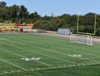 A green sports field with yard lines, a soccer goal, and a football goalpost in the background. Trees and construction equipment are visible beyond the boundary of the field under a clear blue sky.